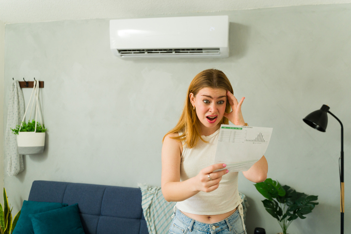Stressed woman looking at expensive electricity bill while standing in living room with air conditioning unit